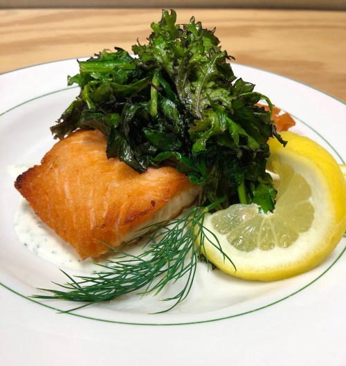 A plate of salmon topped with sautéed greens, served with a lemon wedge and dill on a white dish.