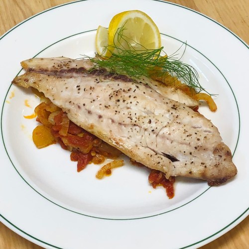 A plated fish fillet garnished with lemon and herbs, served on a bed of sautéed vegetables.