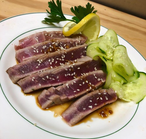 Sliced seared tuna with sesame seeds, served with cucumber slices and a lemon wedge on a white plate.