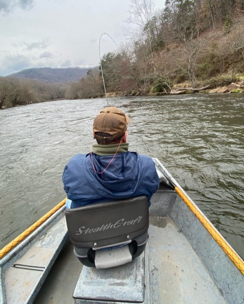 A person fishing from a small boat on a river, with trees and mountains in the background.