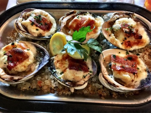 A platter of baked oysters topped with cheese, bacon, and herbs, served with a lemon wedge on a bed of ice.