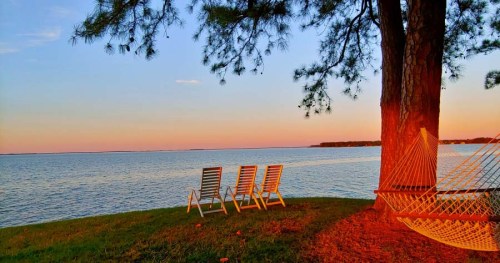 A serene lakeside scene at sunset, featuring a hammock and lounge chairs under a tree.
