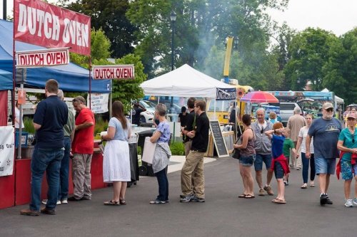 A busy outdoor market scene with people lined up at food stalls, enjoying a sunny day. Tents and food trucks in the background.