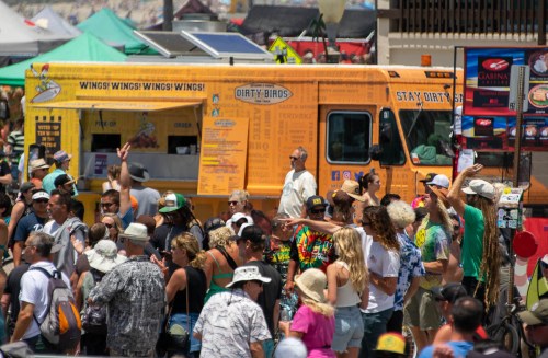 A busy outdoor scene with a vibrant food truck and a crowd of people enjoying a sunny day at a festival.