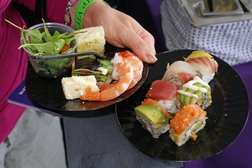 A hand holds two black plates with sushi, shrimp, salad, and a small piece of bread.