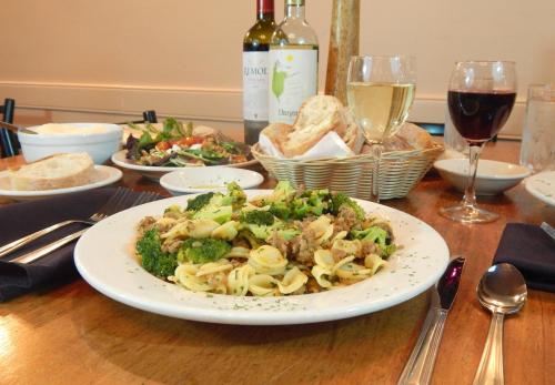 A plate of pasta with broccoli and sausage, surrounded by wine, salad, bread, and other dishes on a wooden table.