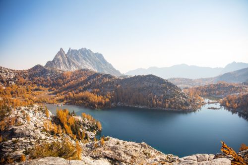 A scenic view of a mountain landscape with a lake, surrounded by autumn foliage and distant peaks under a clear sky.