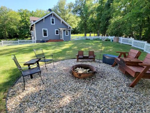 A cozy backyard with a fire pit surrounded by chairs, gravel, and a charming house in the background. Lush greenery surrounds.