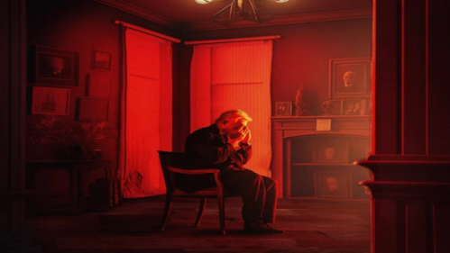 A man sits in a dimly lit, red-toned room, holding his head in his hands, surrounded by framed portraits.