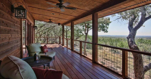 A wooden porch with comfortable seating, overlooking a scenic view of trees and hills under a clear blue sky.