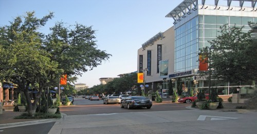 A bustling street scene with trees, shops, and cars under a clear blue sky. Colorful banners hang along the buildings.