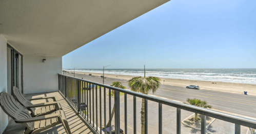 View from a balcony overlooking a beach, with palm trees and a road beside the ocean under a clear blue sky.