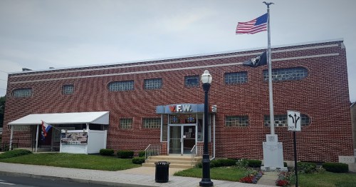 Exterior of a V.F.W. building with an American flag, a tent, and a memorial in front. Green grass and sidewalk visible.