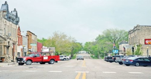 A quiet street scene featuring historic buildings, a red truck, and trees lining the road under a cloudy sky.