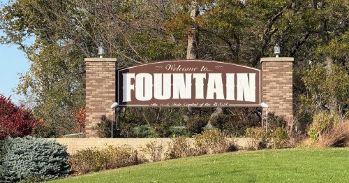 The Fountain, Minnesota welcome sign surrounded by colorful bushes and autumn trees on a clear day.