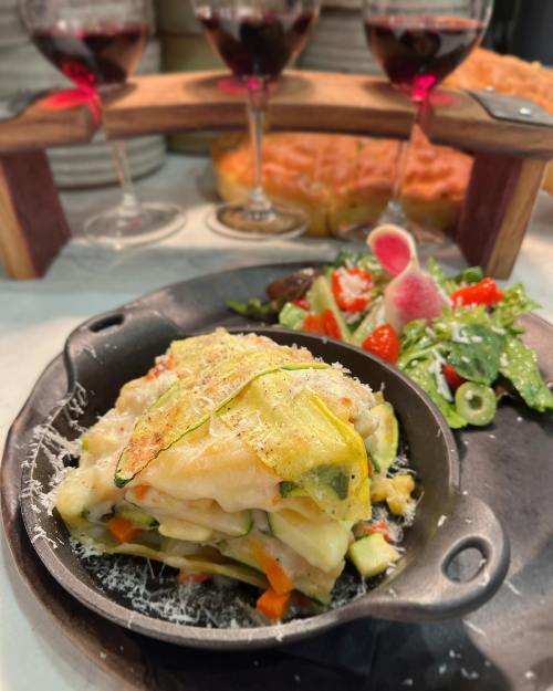 A serving of vegetable lasagna in a black dish, garnished with cheese, alongside a fresh salad and glasses of red wine.