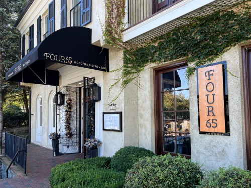 Exterior of FOUR65 Woodfire Bistro & Bar, featuring a black awning and a sign, surrounded by greenery and brick pathway.