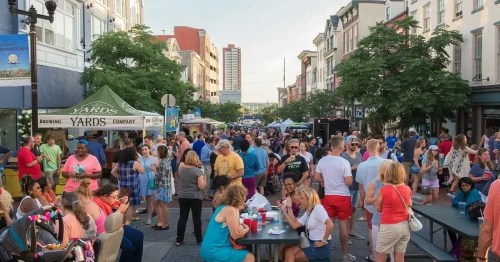 A lively street festival with crowds of people, food stalls, and tents under trees in an urban setting.