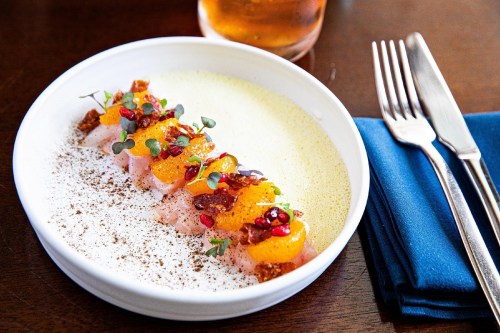 A white plate with colorful sushi topped with citrus, pomegranate, and herbs, served with a frothy sauce.