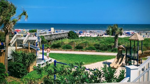 A sunny beach scene with playground equipment, palm trees, and people enjoying the ocean in the background.