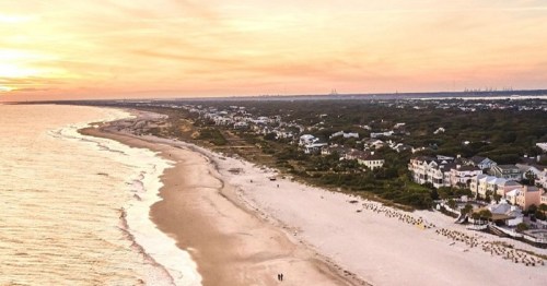 Aerial view of a beach at sunset, with a shoreline lined by houses and a calm ocean.