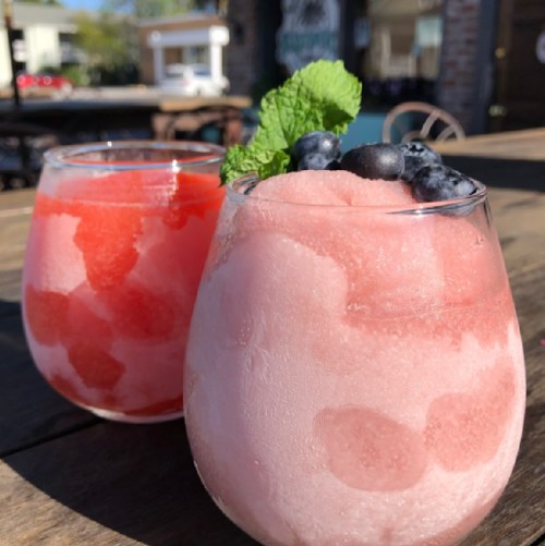 Two colorful frozen drinks in glasses, one red and one pink, garnished with mint and blueberries, on a wooden table.