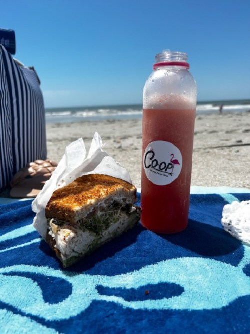 A beach scene featuring a sandwich and a drink on a blue towel, with the ocean in the background.