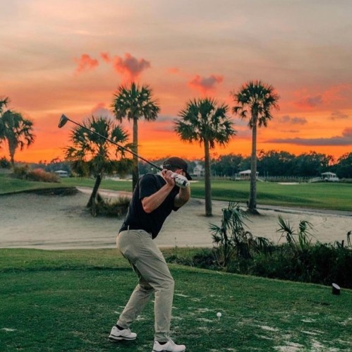 A golfer swings a club at sunset, surrounded by palm trees and a scenic landscape.