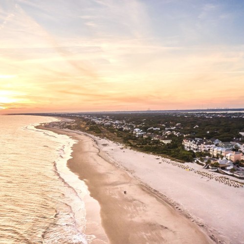 Aerial view of a sandy beach at sunset, with gentle waves and a coastal town in the background.