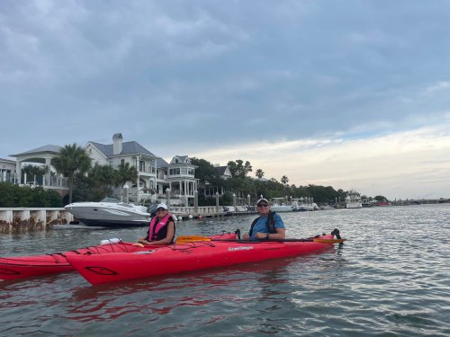 Two people in a red kayak on calm water, with large houses and boats in the background under a cloudy sky.