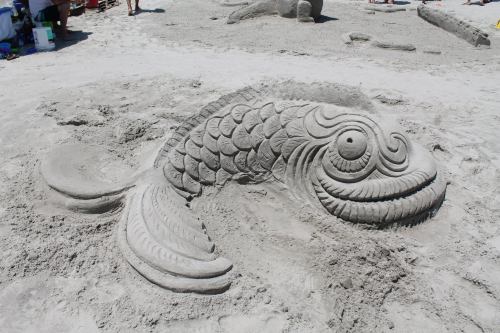 A detailed sand sculpture of a fish with intricate scales, set on a sandy beach.