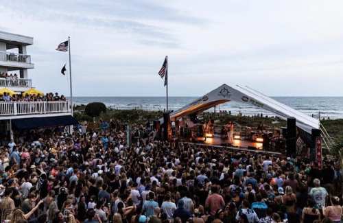A large crowd enjoys a live music performance on a beachside stage, with the ocean and flags in the background.