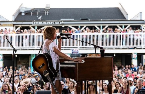 A musician performs on stage with a guitar and piano, while a large crowd enjoys the concert.