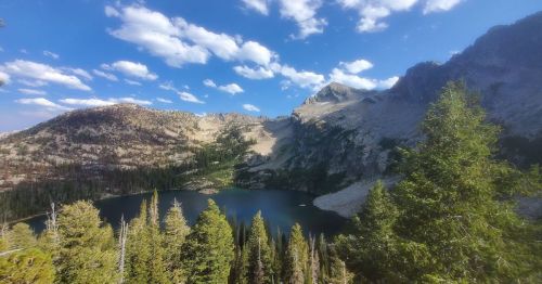 A scenic view of a mountain lake surrounded by trees and rugged mountains under a blue sky with scattered clouds.
