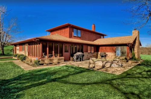 A red two-story house with a patio, surrounded by green grass and trees under a clear blue sky.