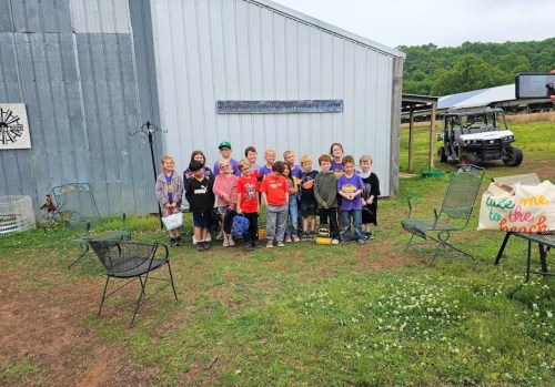 A group of children stands outside a barn, smiling and posing for a photo on a cloudy day.