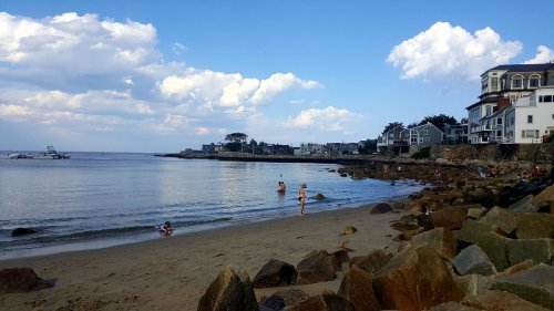 A serene beach scene with people wading in the water, rocky shore, and houses along the coastline under a blue sky.