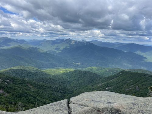 A panoramic view of lush green mountains under a cloudy sky, with rocky foreground and distant peaks.