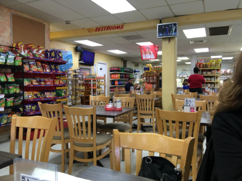 A cozy café with wooden tables, snack shelves, and a TV, featuring a few customers and a restroom sign.