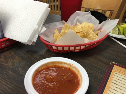 A bowl of salsa next to a basket of tortilla chips on a wooden table, with a drink and napkins in the background.