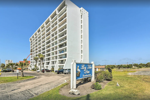 A white multi-story building with a sign reading "By The Sea" and palm trees in the foreground under a clear blue sky.