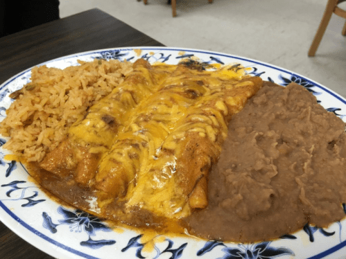 A plate of enchiladas topped with cheese, served with rice and refried beans on a decorative blue and white dish.