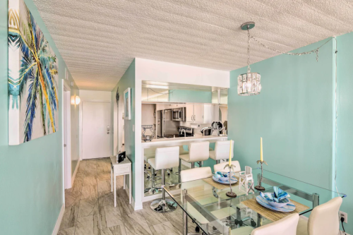 Bright, modern dining area with a glass table, white chairs, and turquoise walls, leading to a kitchen with bar seating.