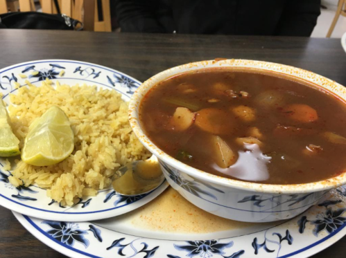 A bowl of rich soup with vegetables and meat, served with a side of yellow rice and lime wedges on a decorative plate.