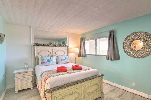 Cozy bedroom with a light blue wall, floral bedding, and decorative mirror, featuring a bedside table and natural light.