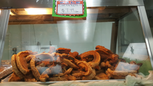 A display case filled with crispy pork cracklings, labeled "Chicharrón" for sale at $14.00.