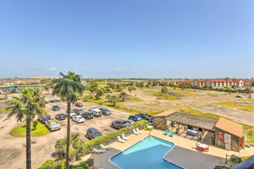 View of a pool area with palm trees, parked cars, and a clear sky, overlooking a vacant lot and nearby buildings.