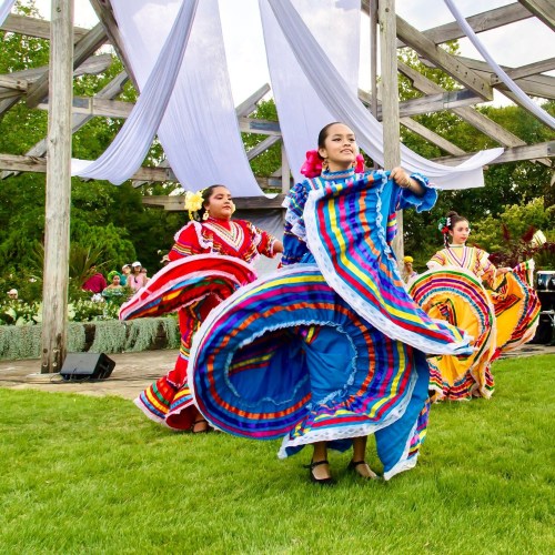 Two dancers in colorful traditional dresses perform on a grassy outdoor stage, surrounded by greenery and decorations.