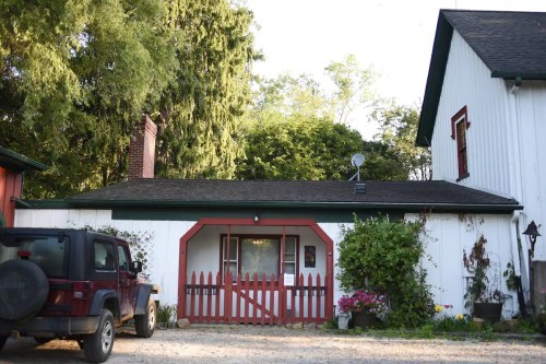 A quaint white house with red accents, featuring a small fenced area and a parked black Jeep in front.
