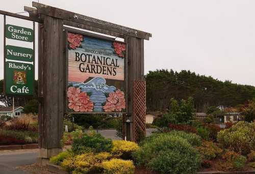 Sign for Mendocino Coast Botanical Gardens, featuring colorful flowers and nearby greenery, with a café and nursery.